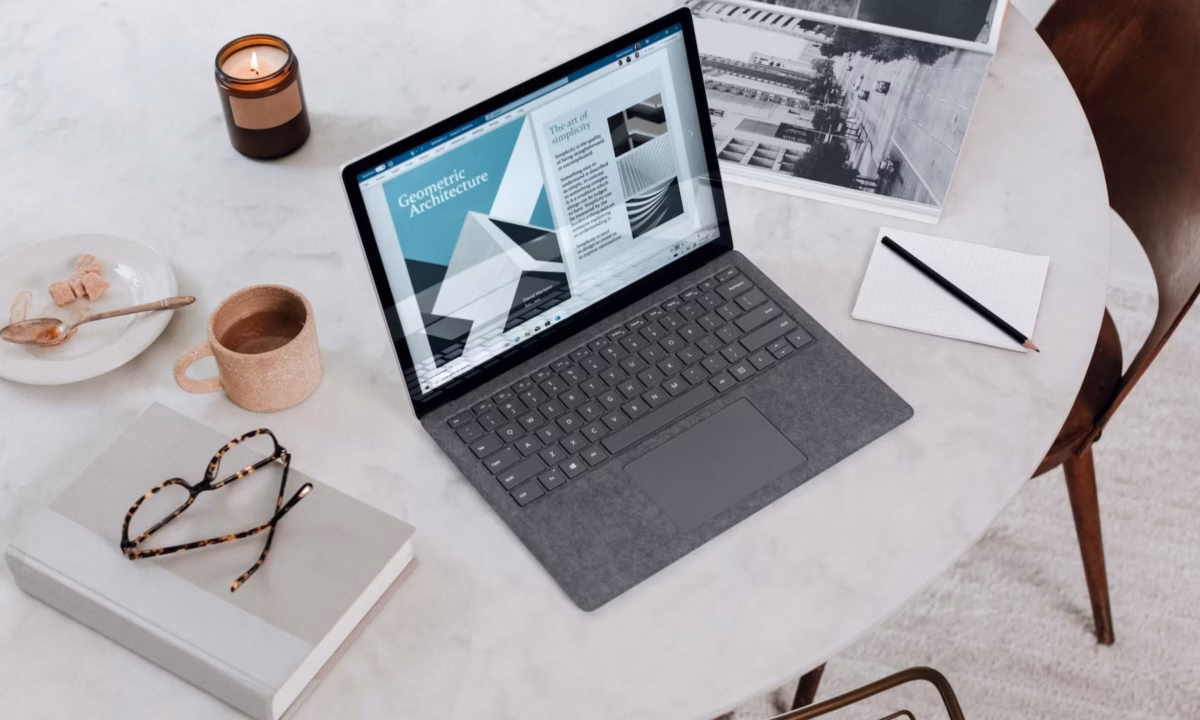 A laptop, notebook, glasses, notepad, pen, coffee, and candle are on top of a table.