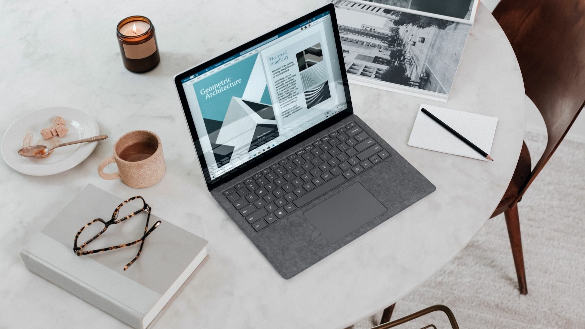 A laptop, notebook, glasses, notepad, pen, coffee, and candle are on top of a table.