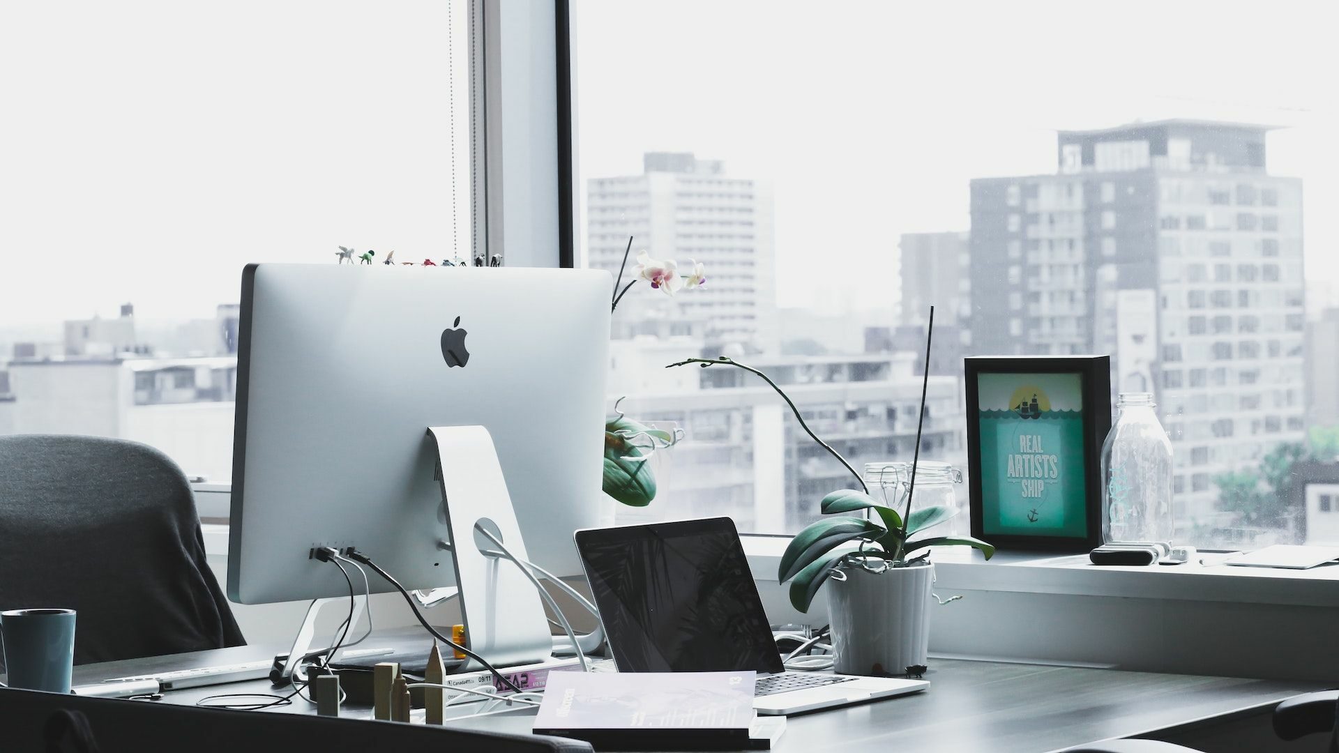 A workspace with a Mac, plants, and city buildings in the background.