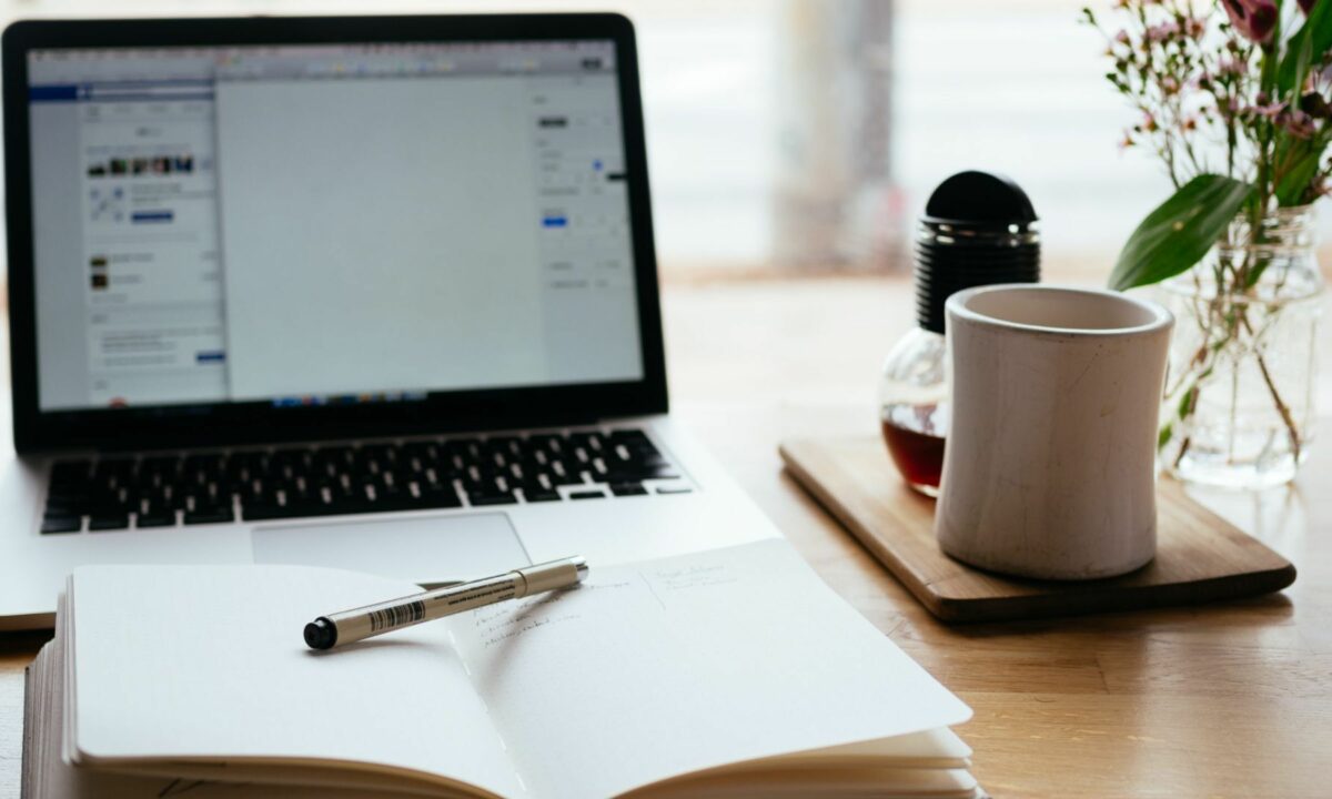 A laptop, a pen on top of an open notebook, a cup of coffee, and a plant.