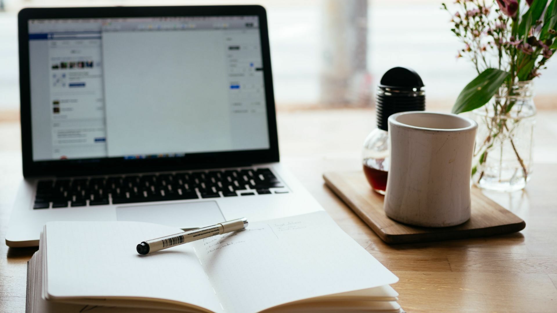 A laptop, a pen on top of an open notebook, a cup of coffee, and a plant.