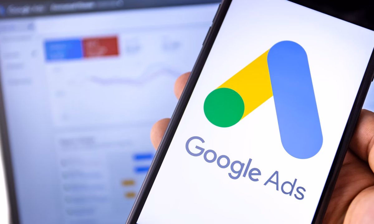 A mobile phone with the Google Ads account logo and a laptop in the background with the Google Ads for real estate dashboard on the screen.