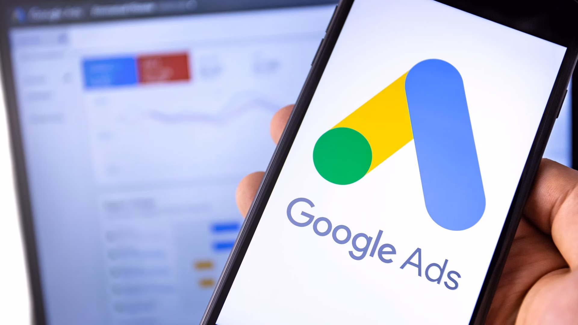 A mobile phone with the Google Ads account logo and a laptop in the background with the Google Ads for real estate dashboard on the screen.