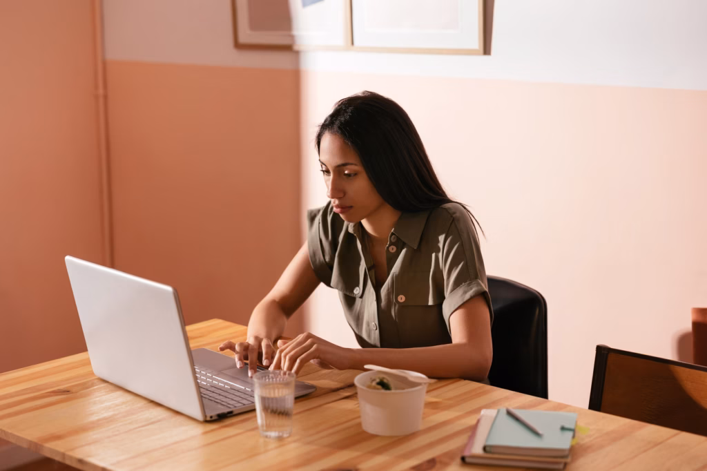 Focused female freelancer browsing netbook and on remote project while sitting at table with takeaway food and glass of water