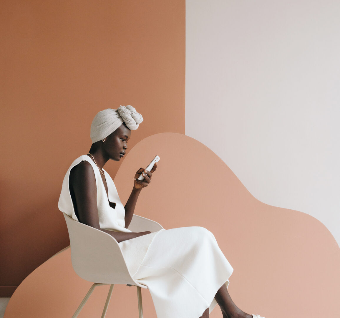 Side view of confident young black female model in trendy total white outfit and traditional turban sitting on chair and messaging on smartphone on two color background