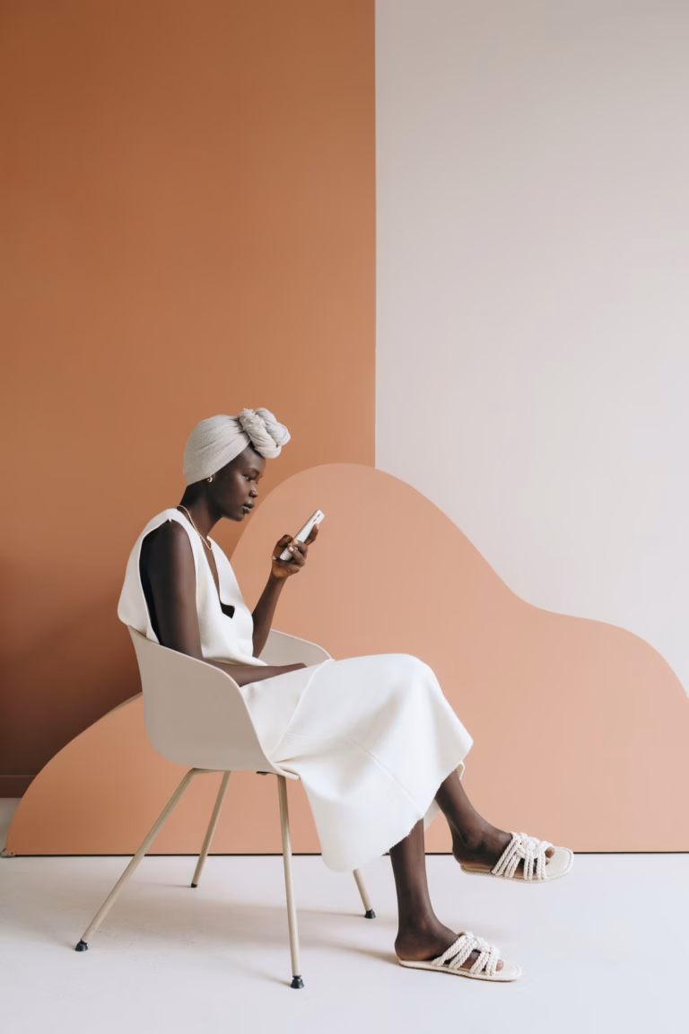 Side view of confident young black female model in trendy total white outfit and traditional turban sitting on chair and messaging on smartphone on two color background