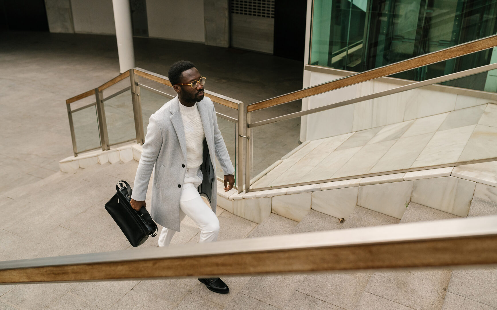 From above of serious African American male entrepreneur in elegant outfit with briefcase walking upstairs of modern business center in city