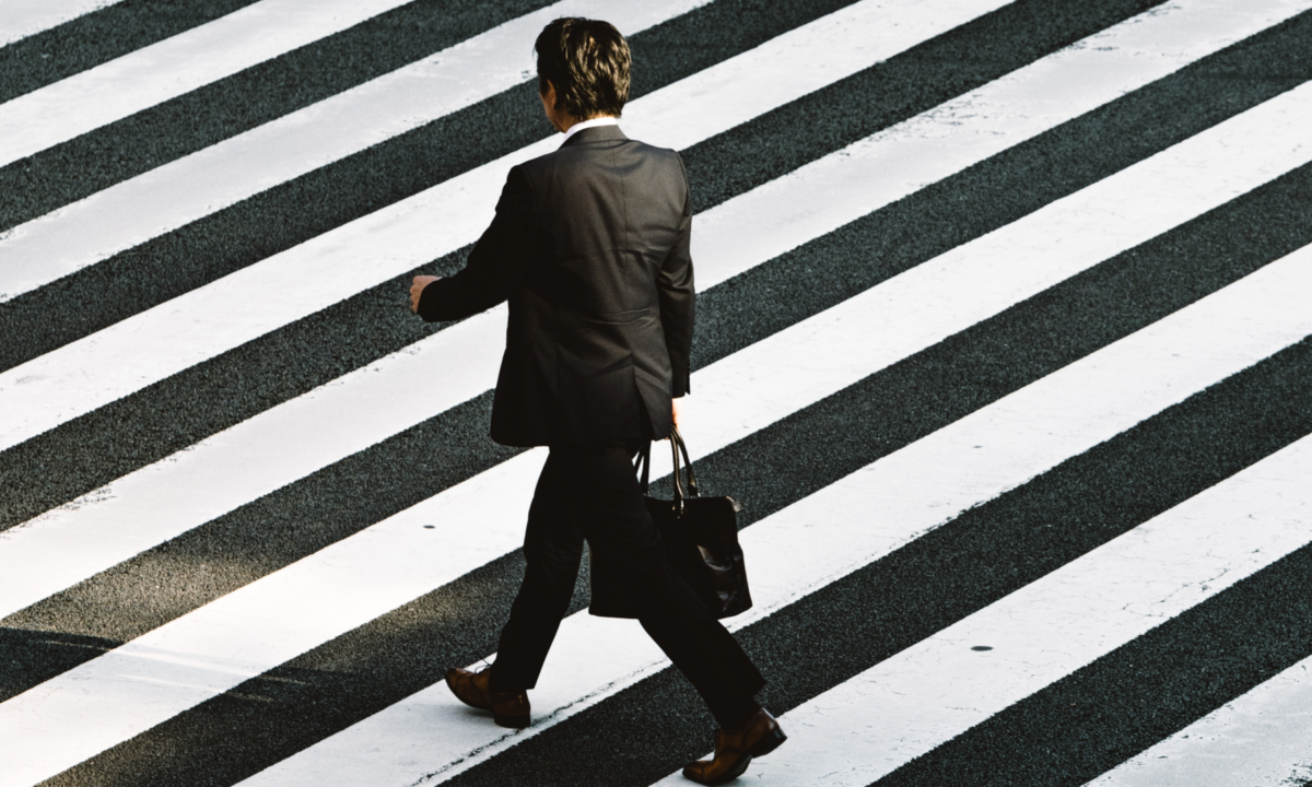 A male real estate professional in a black suit walking with briefcase and stands out at a road crossing.