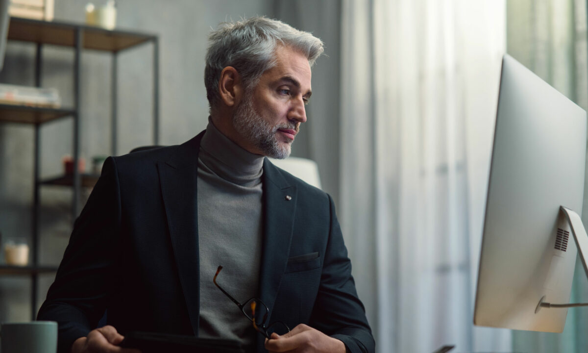 Older male real estate agent sitting at desk looking at the computer screen and auditing his current branding to attract more business.