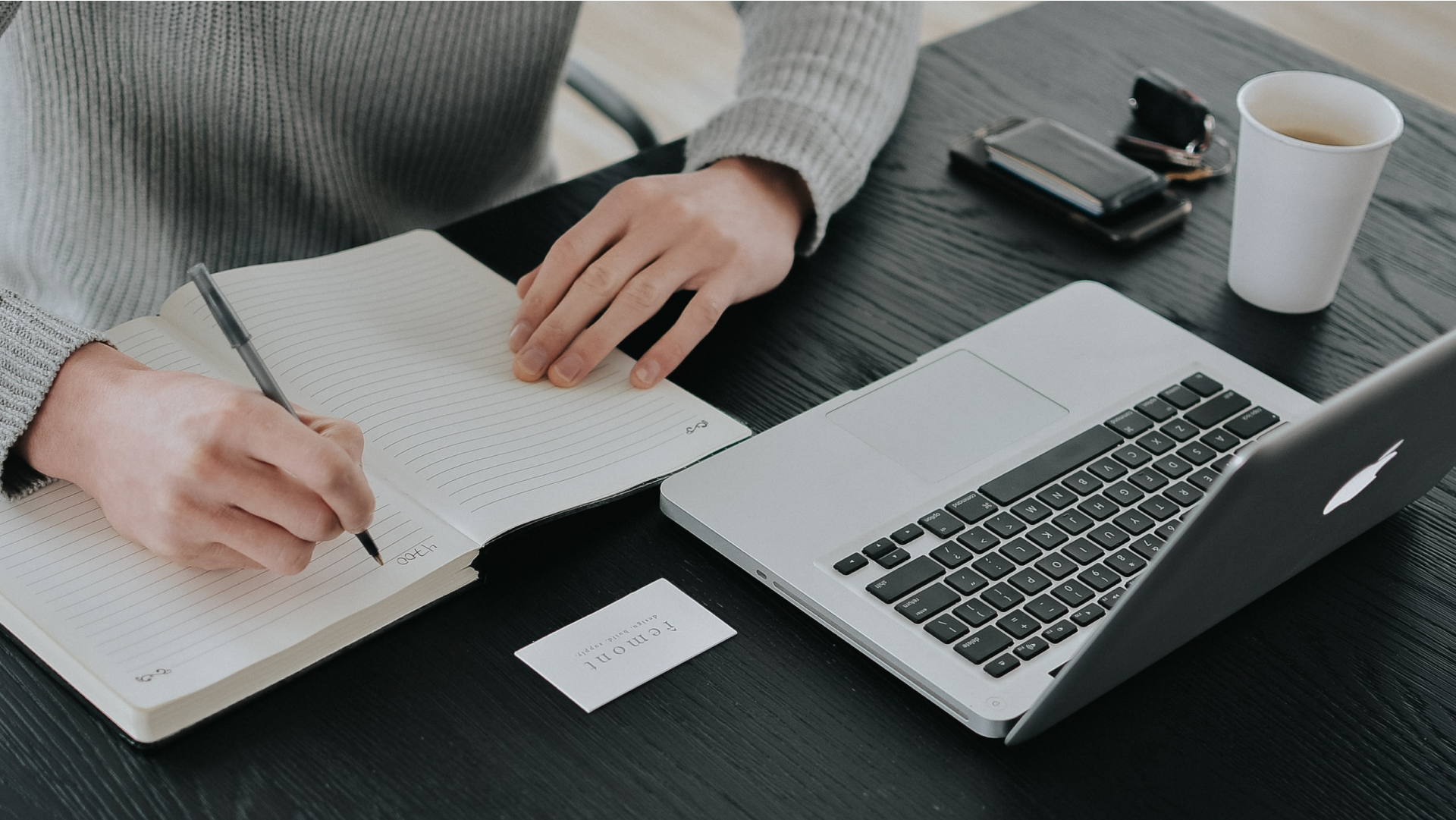 Image of person sitting at desk with laptop open and writing in a notepad thats situated next to a coffee cup