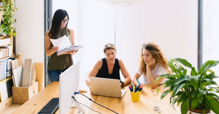 Three real estate agents gather around a table with a laptop and paperwork, working on growing their business