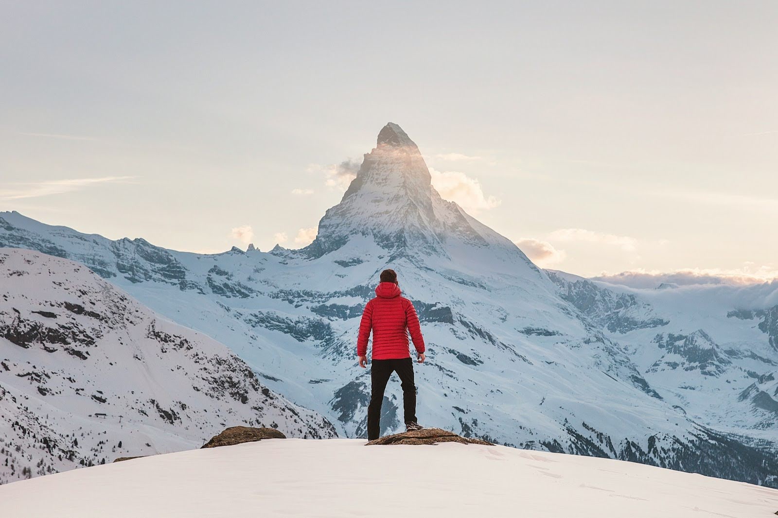A person in a red coat stands at the top of a snowy hill, looking up at a mountaintop that is blocking the sun and shrouded in clouds.