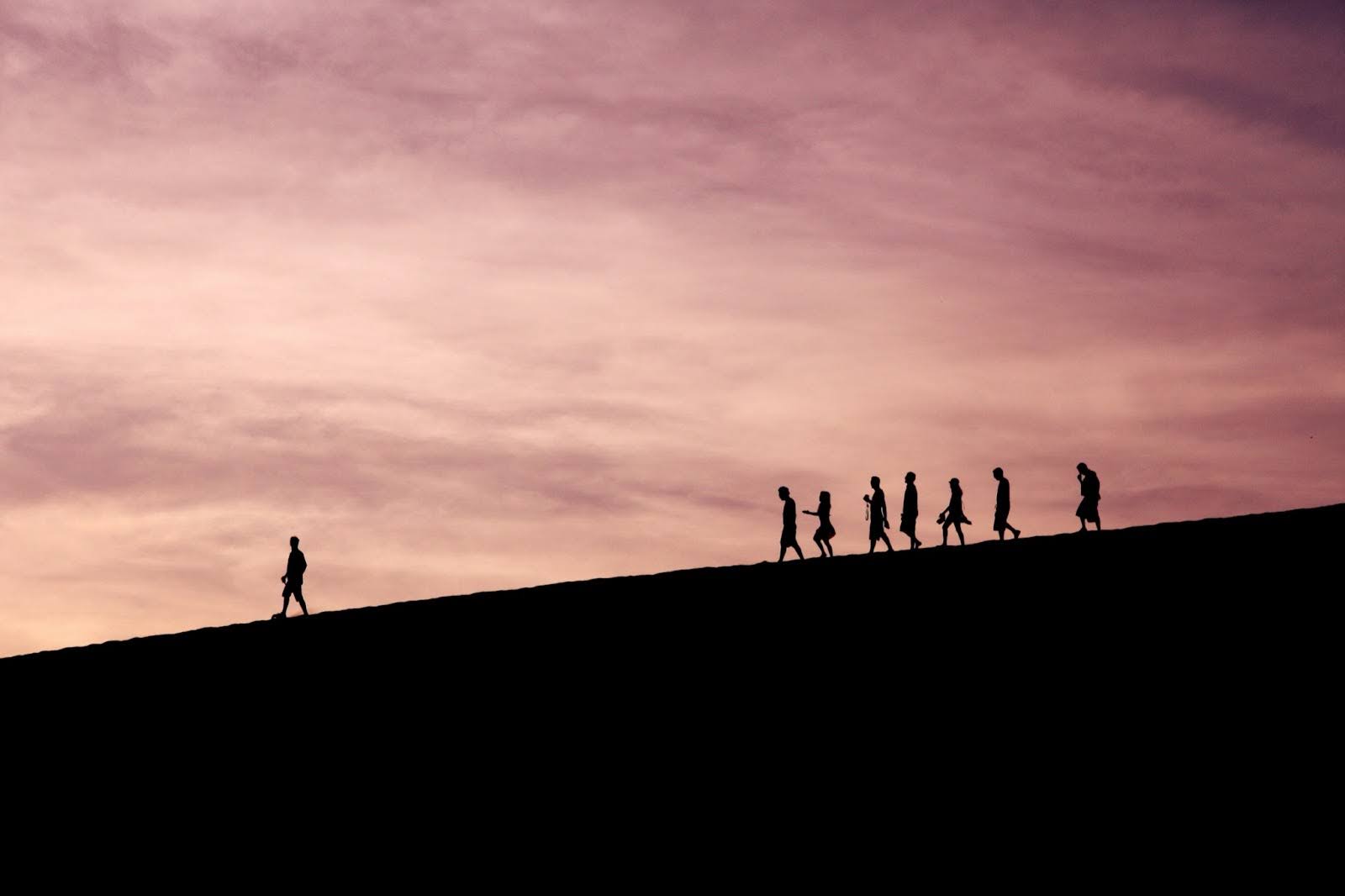 Seven backlit figures walk down a hill, following a leader at the front walking ahead of the group