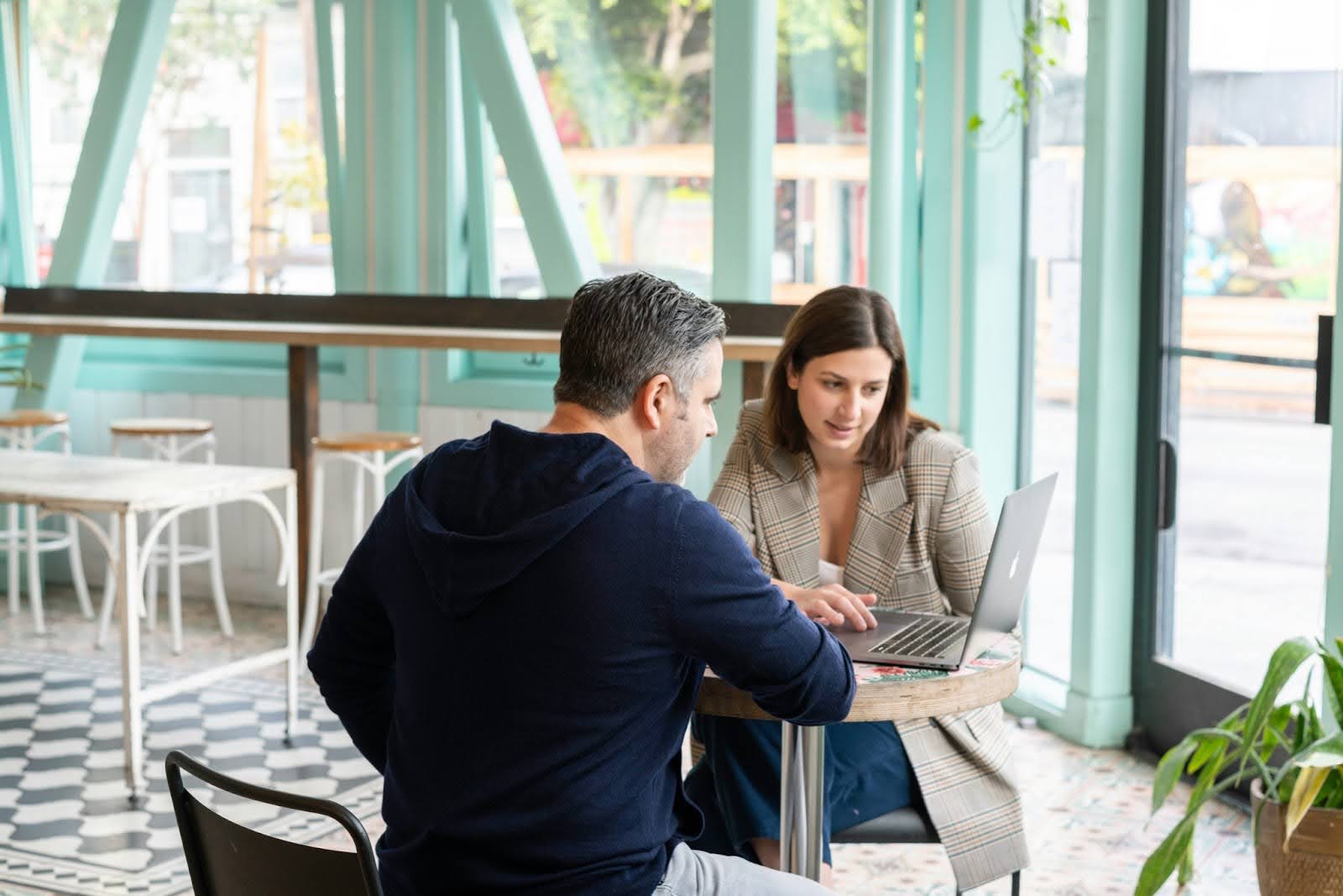 A real estate professional sits at a table in a cafe with teal walls and many windows, demonstrating to her potential client her various sales strategies for his listing on her laptop