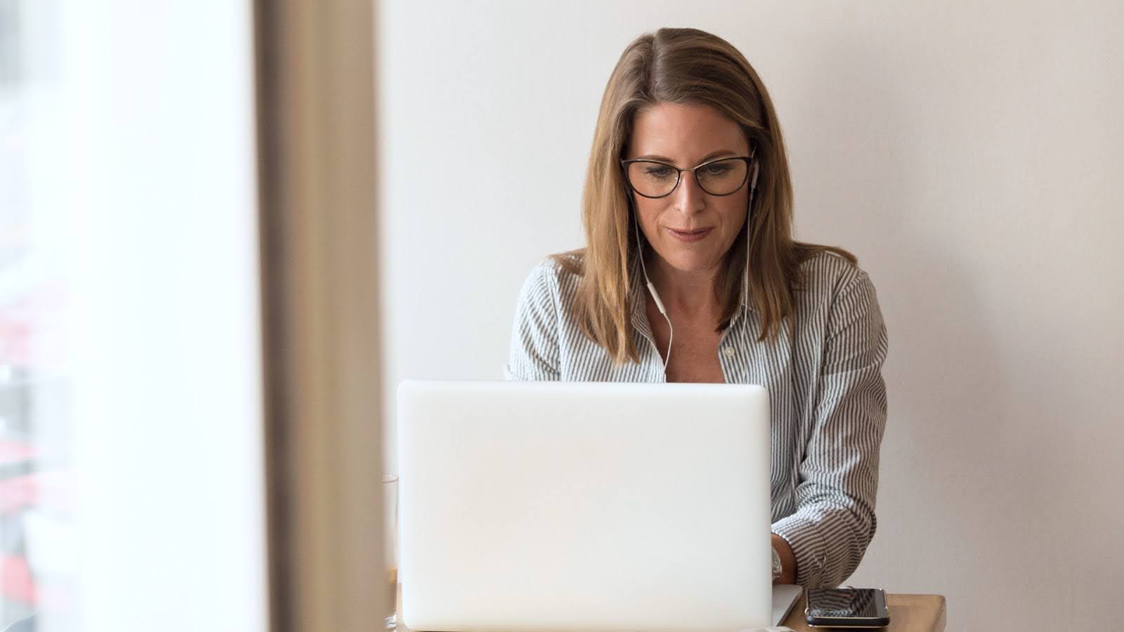 A real estate agent with earbuds in sits at a small desk typing on her laptop with her smartphone next to her