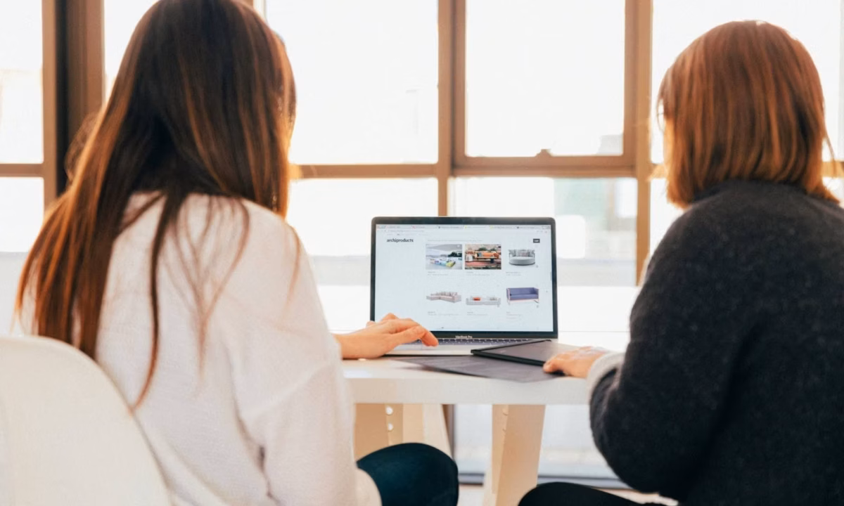Two businesspeople sit in front of a laptop looking at metrics in front of a bright window