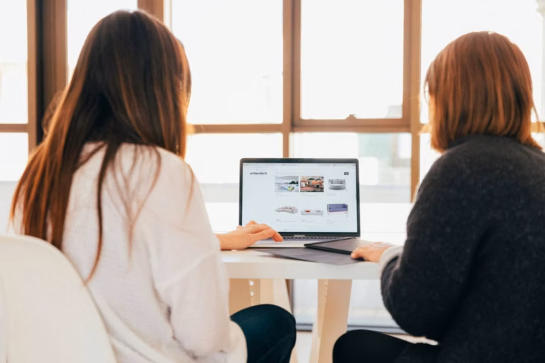 Two businesspeople sit in front of a laptop looking at metrics in front of a bright window