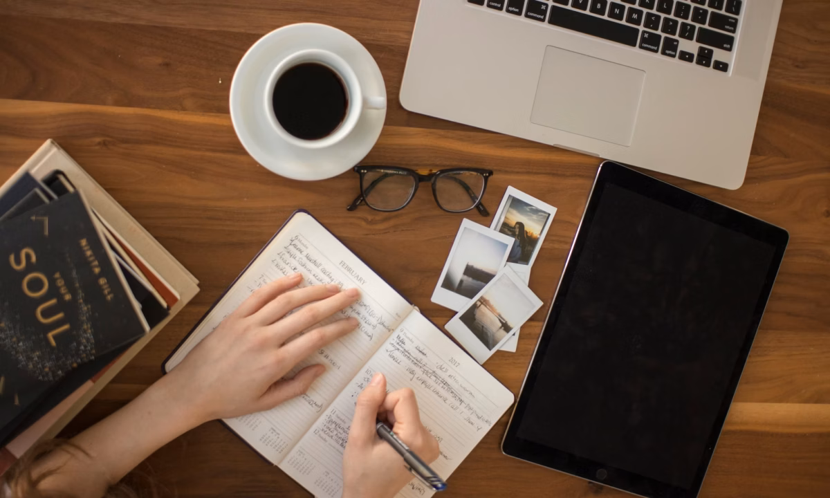 Woman updates calendar in front of cup of black coffee, branding and marketing inspiration photos, laptop and several books on a desk