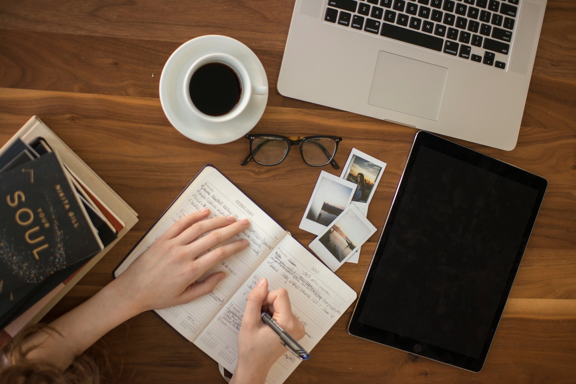 Woman updates calendar in front of cup of black coffee, branding and marketing inspiration photos, laptop and several books on a desk