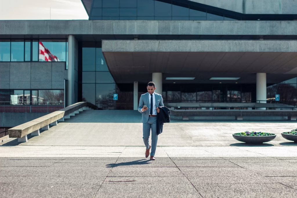 Business man walking toward camera while holding a coat and looking at his phone in front of a modern concrete building
