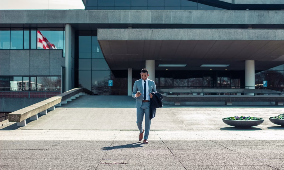 Business man walking toward camera while holding a coat and looking at his phone in front of a modern concrete building