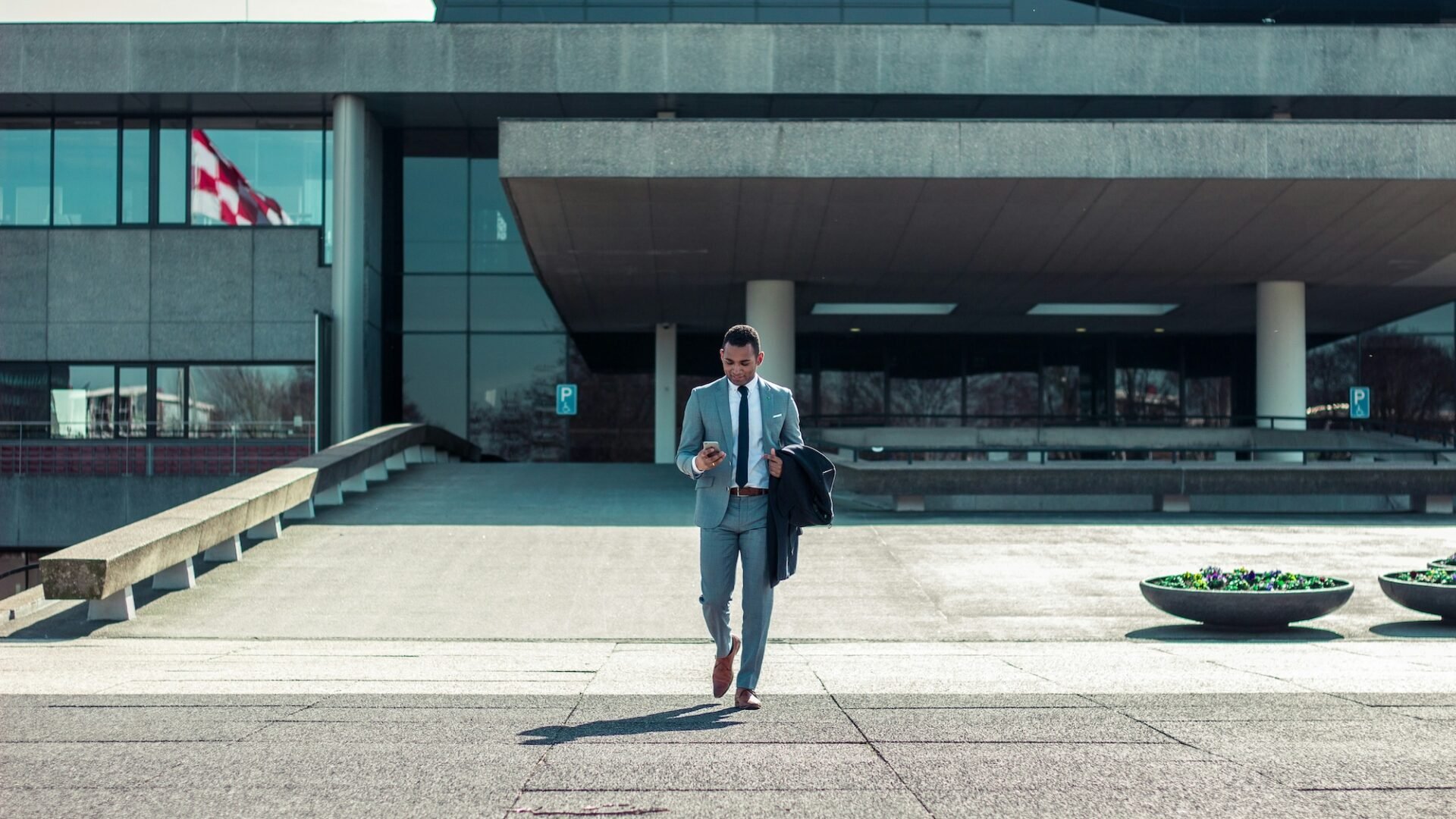 Business man walking toward camera while holding a coat and looking at his phone in front of a modern concrete building