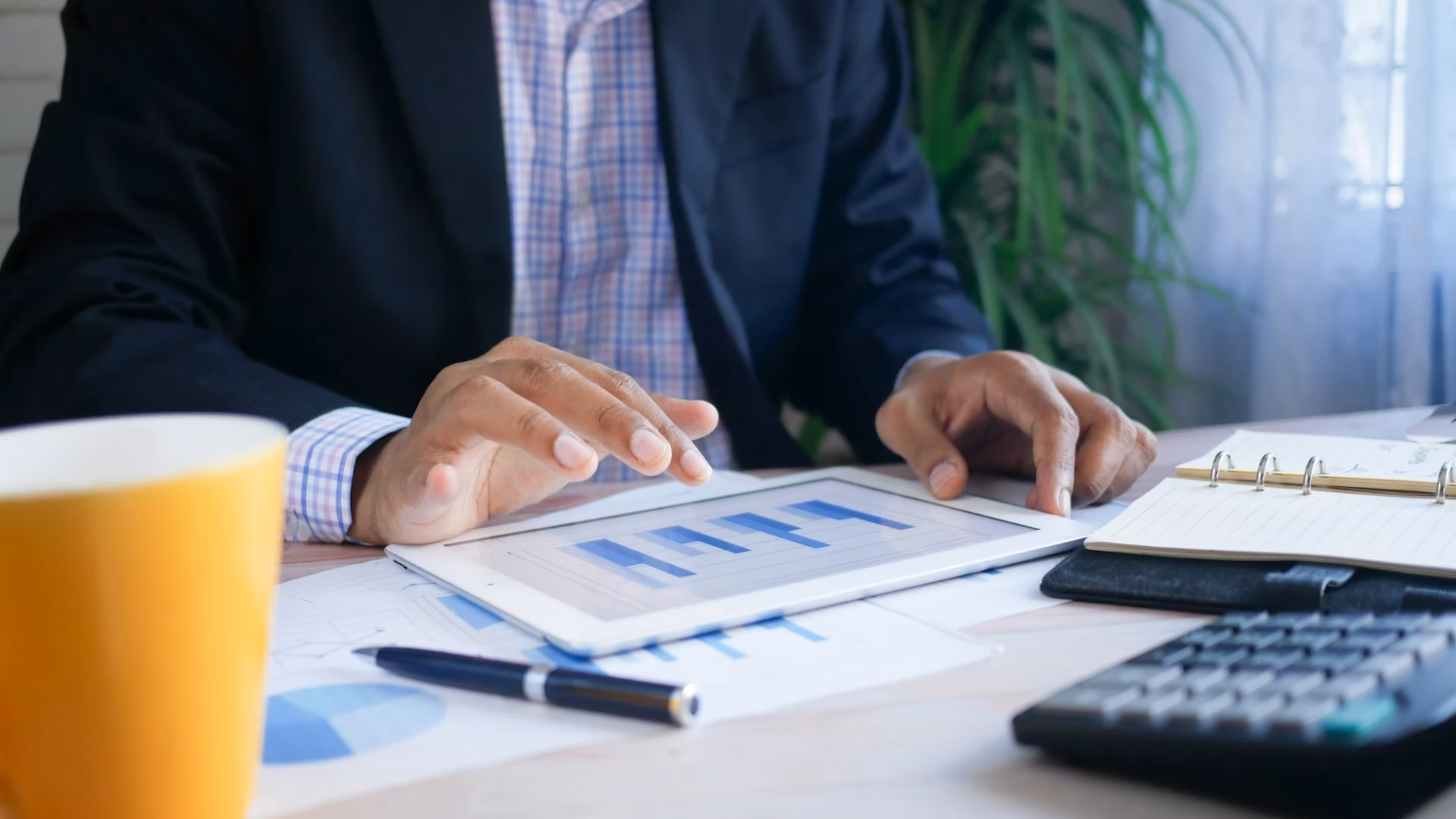 Real estate agent figuring out expenses at a desk with a calculator, tablet and cup of coffee in front of him