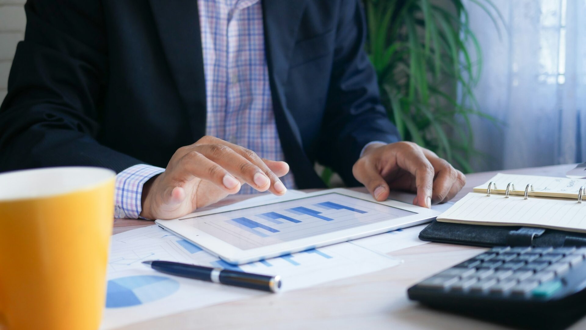 Real estate agent figuring out expenses at a desk with a calculator, tablet and cup of coffee in front of him