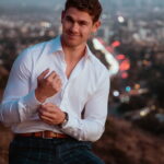 Aaron Grushow stands before LA skyline buttoning his sleeve and smiling at the camera