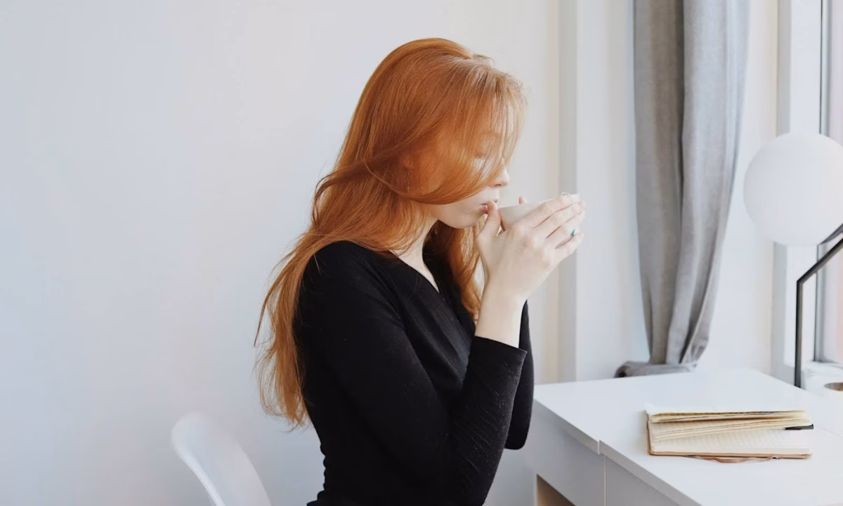 woman sitting at white desk in front of notebook, sipping a cup of tea showing and example of how to invest in yourself