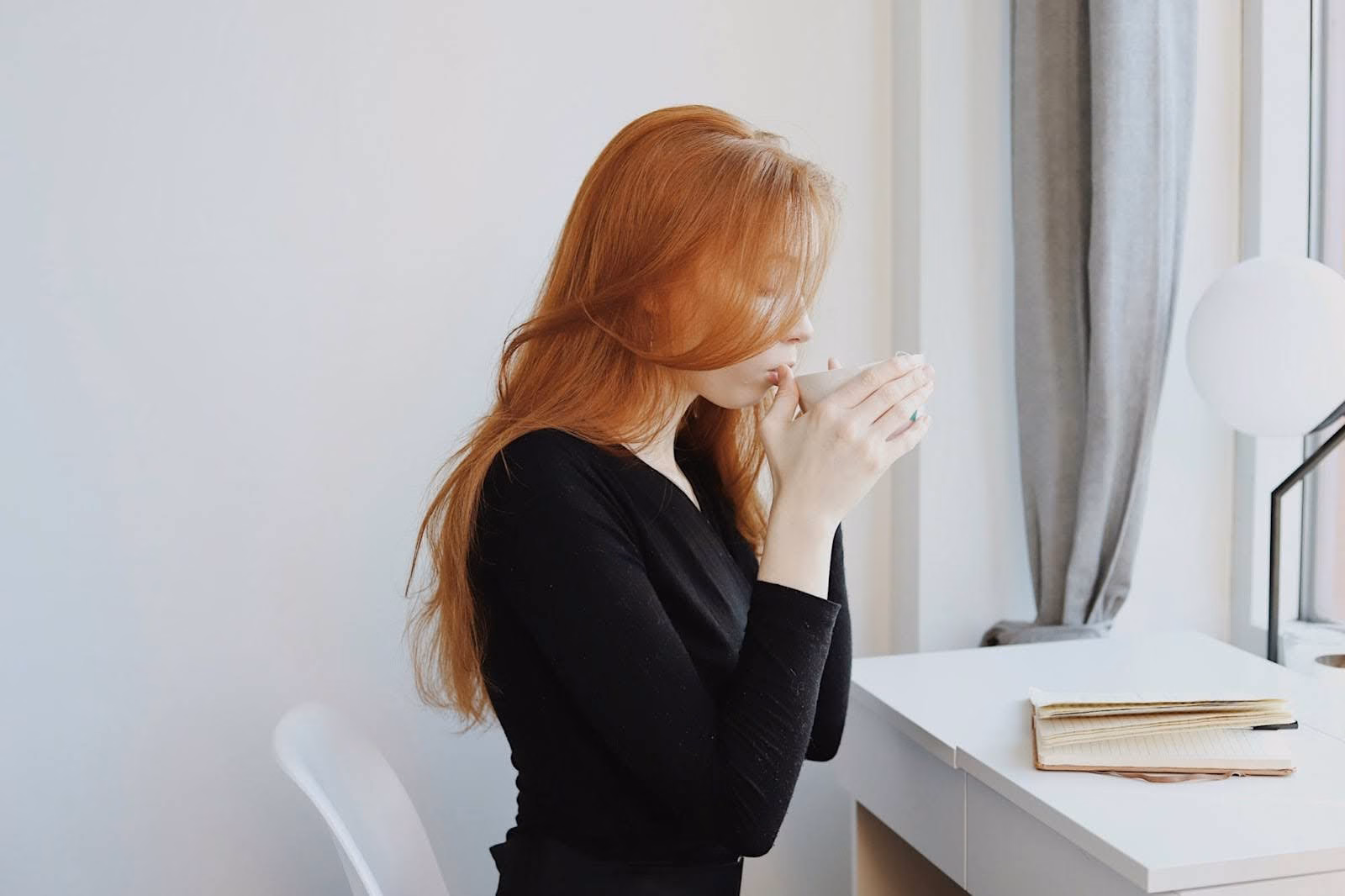 woman sitting at white desk in front of notebook, sipping a cup of tea showing and example of how to invest in yourself