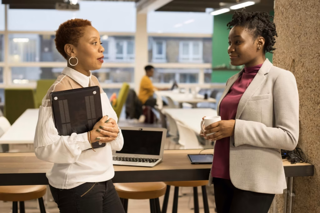 A broker stands in an open office holding a tablet and talking to a real estate agent she is trying to recruit, who is hold a cup of coffee