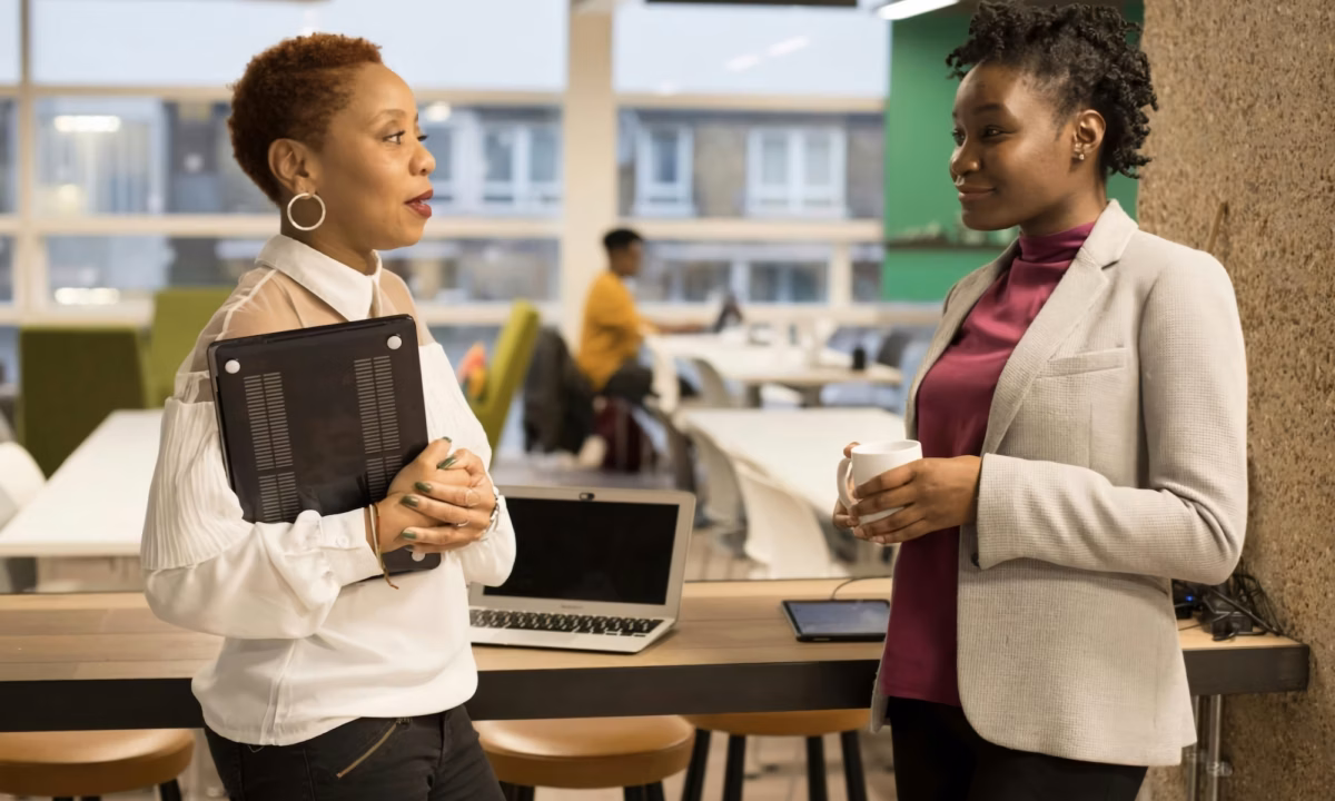 A broker stands in an open office holding a tablet and talking to a real estate agent she is trying to recruit, who is hold a cup of coffee