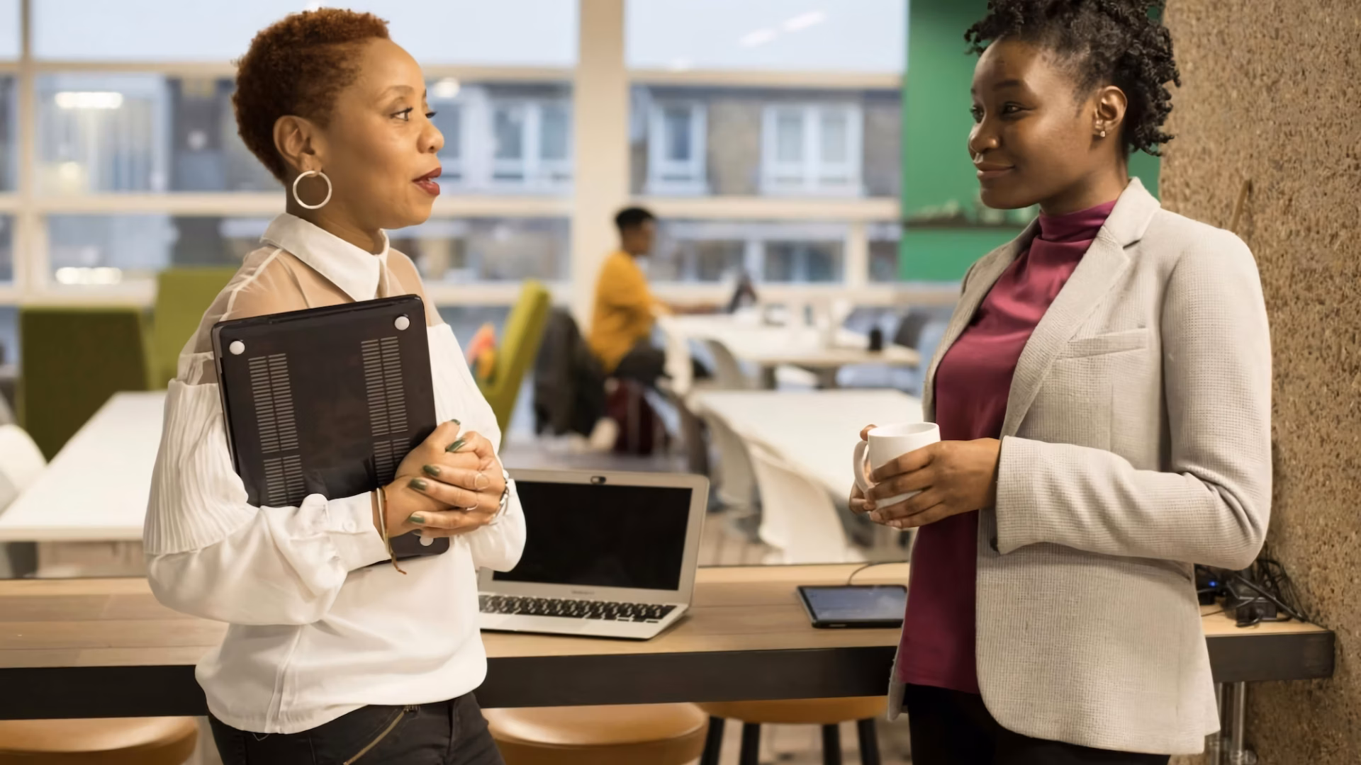 A broker stands in an open office holding a tablet and talking to a real estate agent she is trying to recruit, who is hold a cup of coffee
