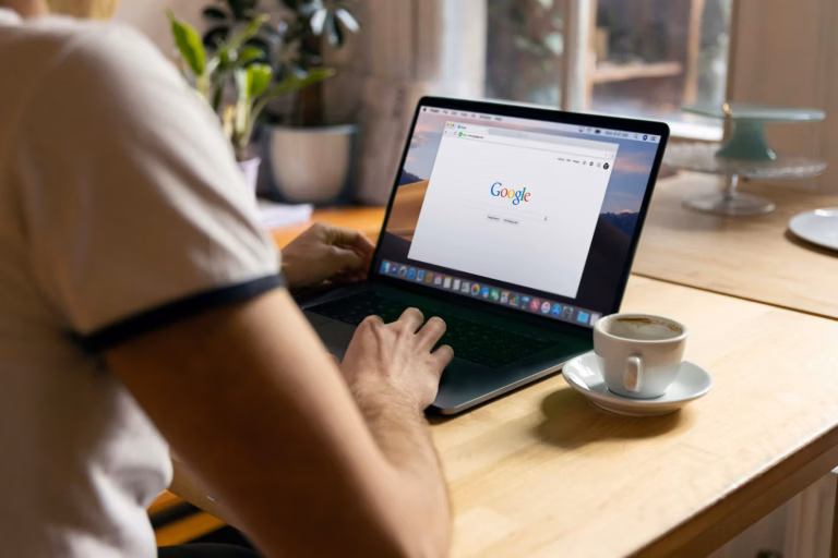 A person sits in front of an empty coffee cup and a laptop with Google's search page open on the screen to illustrate the need to grow your google business profile audience as a real estate agent