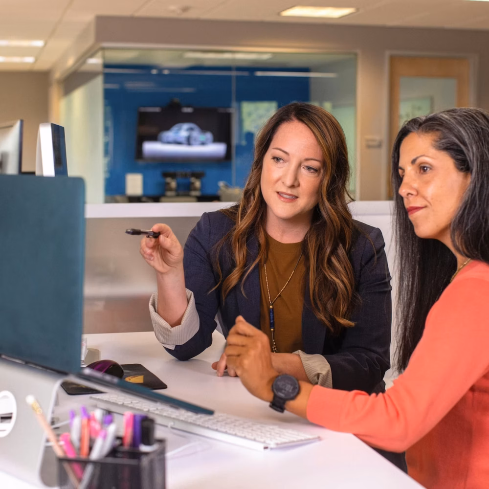 Two women stand at a desk in an office looking at a desktop computer, with one pointing at the screen