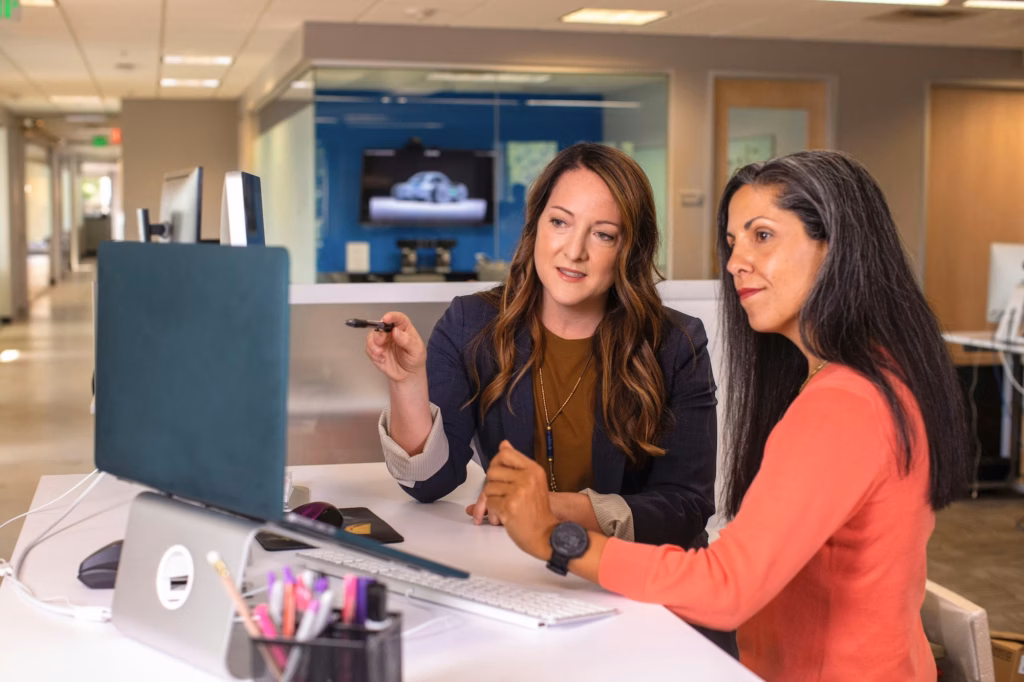 Two women stand at a desk in an office looking at a desktop computer, with one pointing at the screen