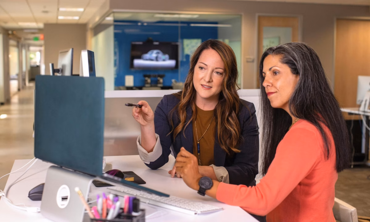 Two women stand at a desk in an office looking at a desktop computer, with one pointing at the screen