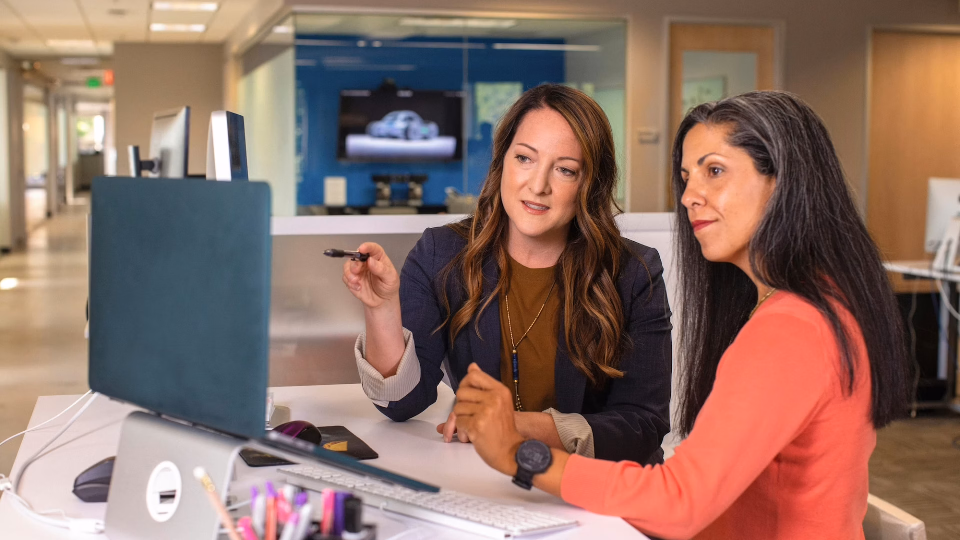 Two women stand at a desk in an office looking at a desktop computer, with one pointing at the screen