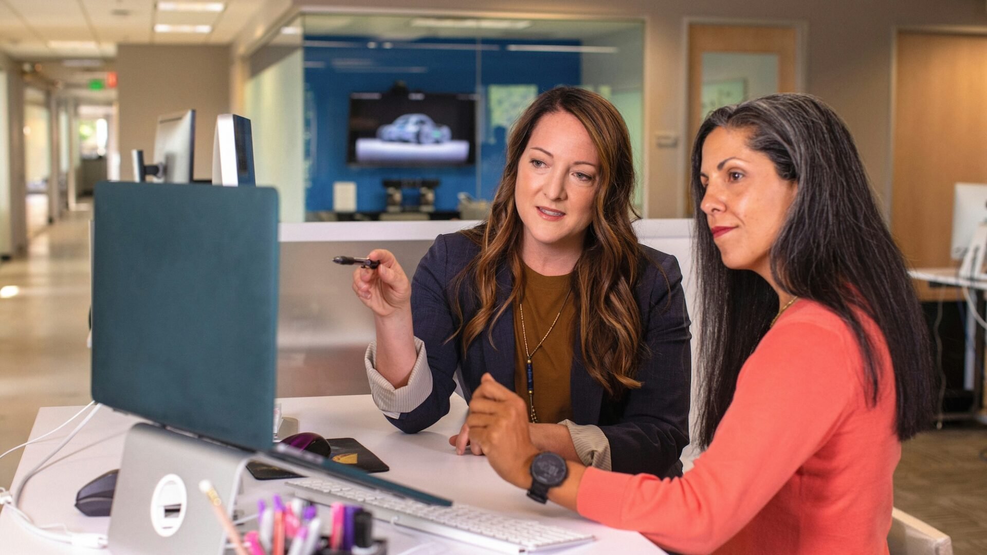 Two women stand at a desk in an office looking at a desktop computer, with one pointing at the screen