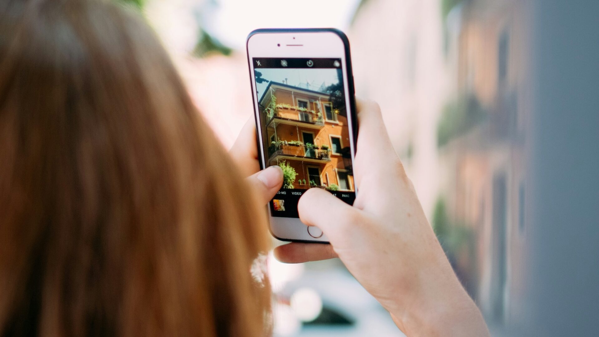 Woman holding up smartphone taking a listing photo of a small condo building for her real estate social media marketing campaign