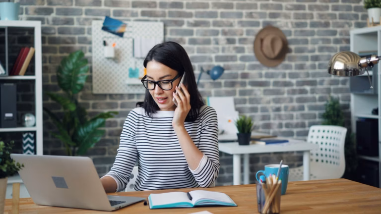 A real estate agent sits at a desk in front of a laptop on her phone, trying out real estate lead conversion ideas to increase business