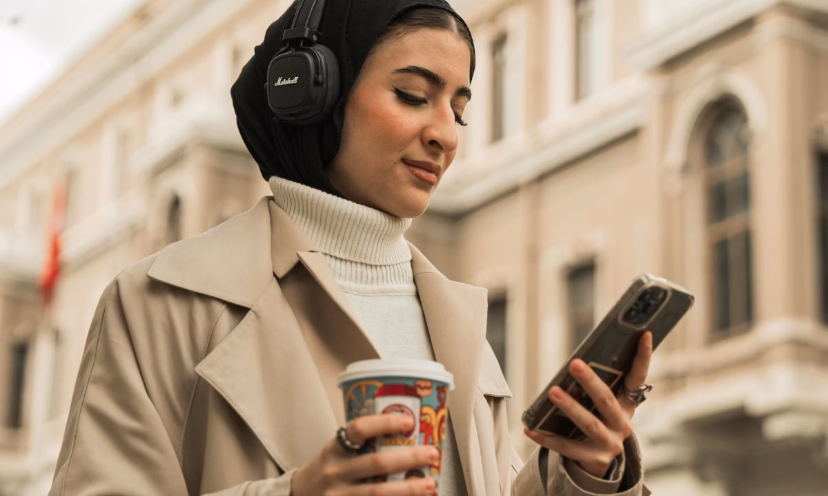 Woman in tan coat walking in city wearing headphones and reading real estate marketing content on a smartphone, emphasizing the need for agents and brokers to optimize for mobile