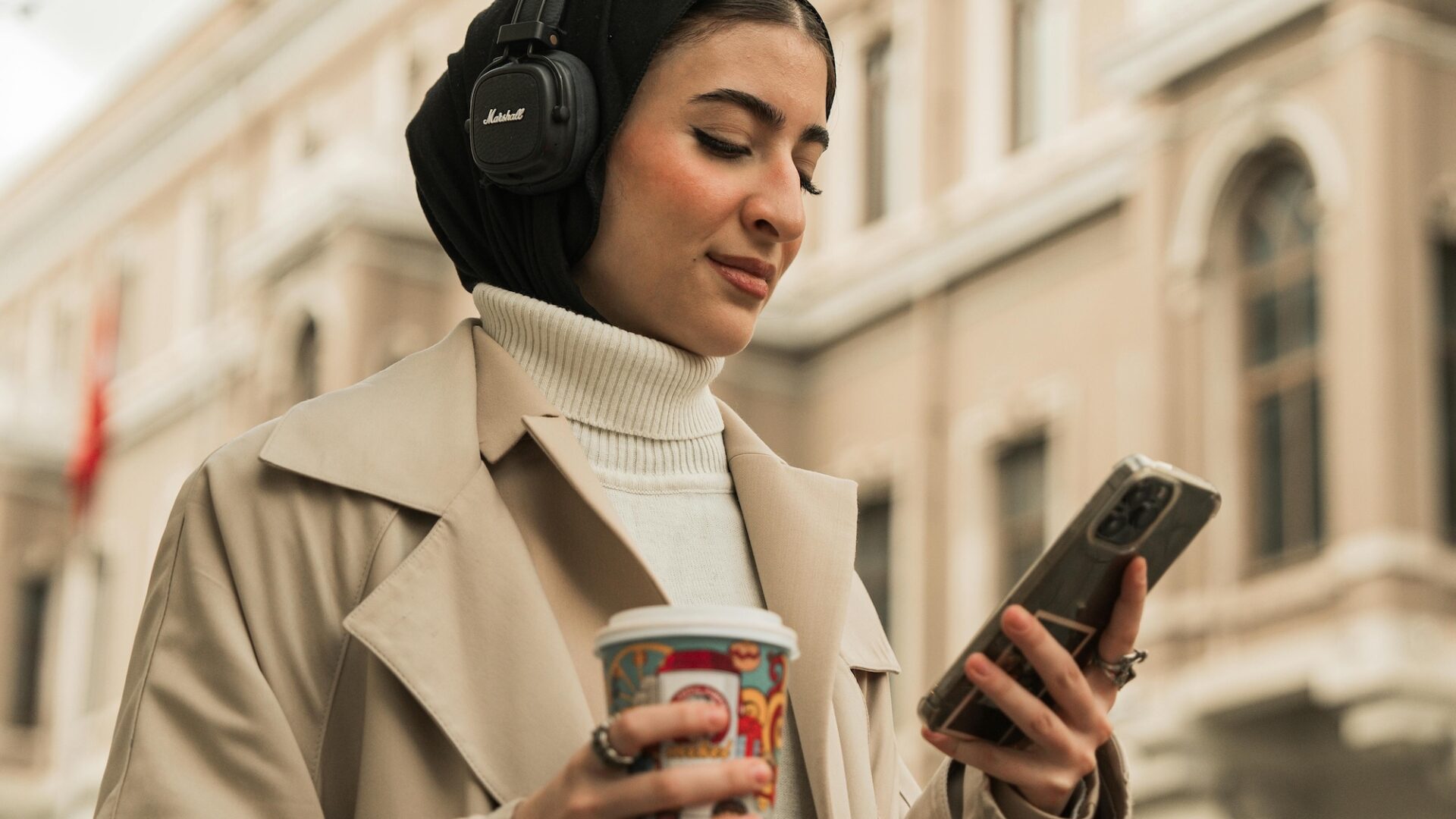 Woman in tan coat walking in city wearing headphones and reading real estate marketing content on a smartphone, emphasizing the need for agents and brokers to optimize for mobile