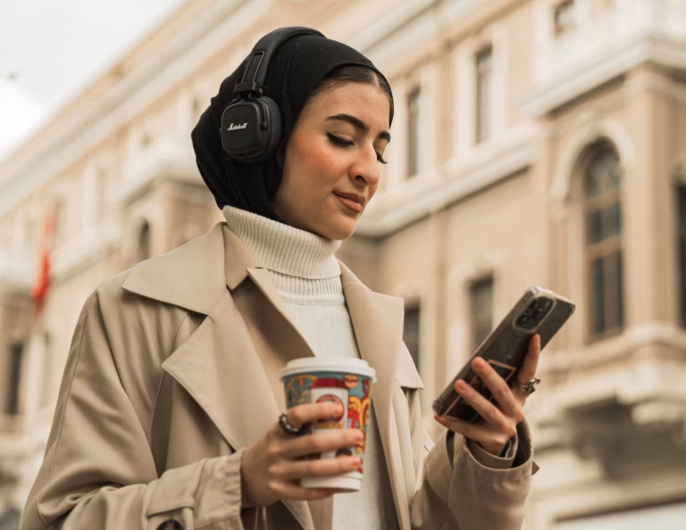 Woman in tan coat walking in city wearing headphones and reading real estate marketing content on a smartphone, emphasizing the need for agents and brokers to optimize for mobile