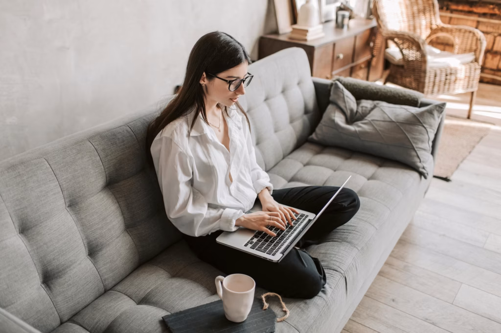 A real estate agent sits crosslegged on a grey couch, typing out a drip email marketing real estate campaign