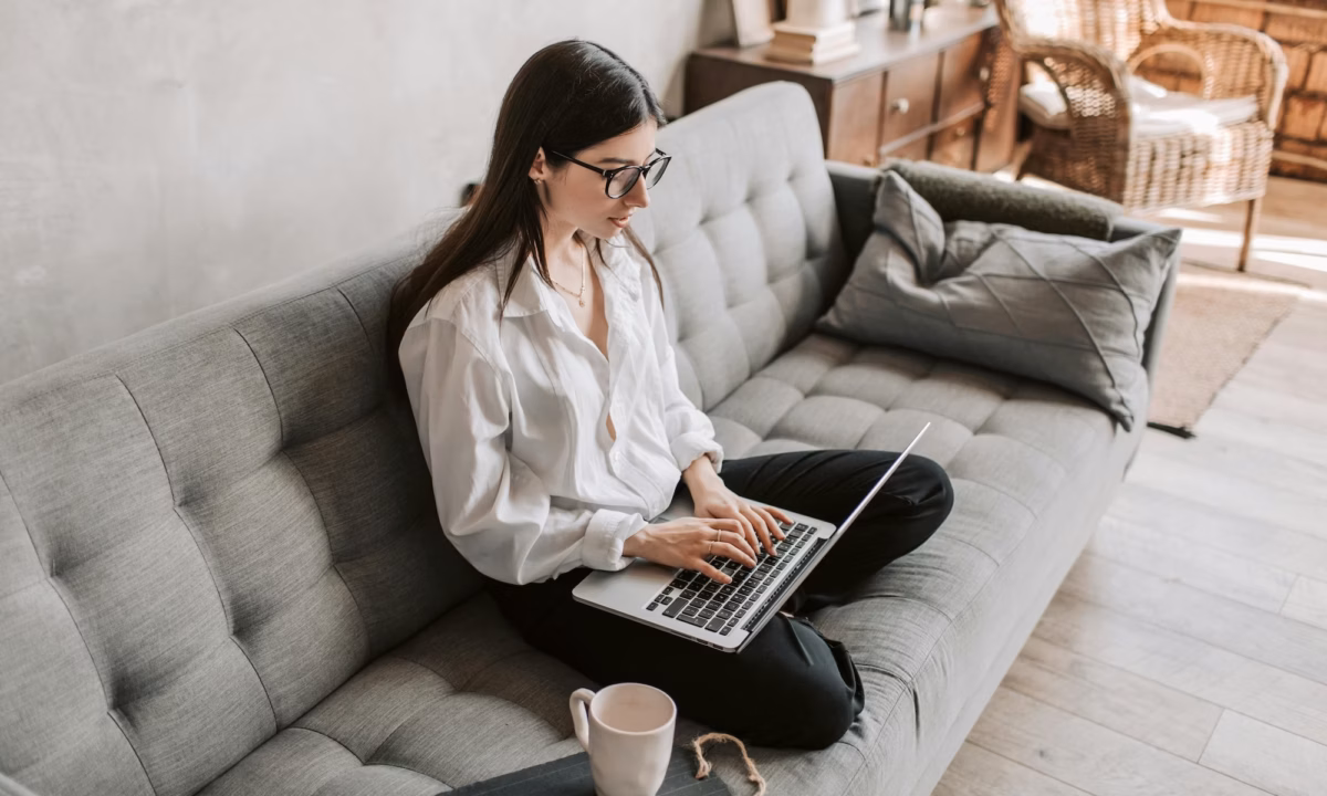 A real estate agent sits crosslegged on a grey couch, typing out a drip email marketing real estate campaign