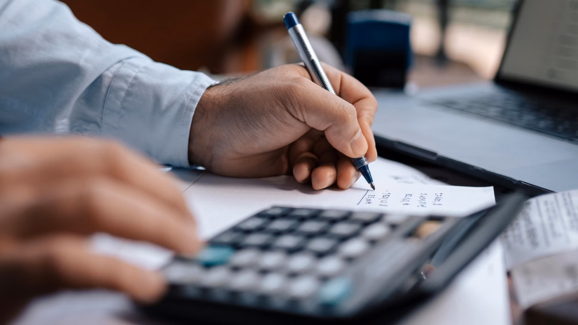 A real estate agent uses a calculator, laptop and pen and paper to figure out their email marketing costs