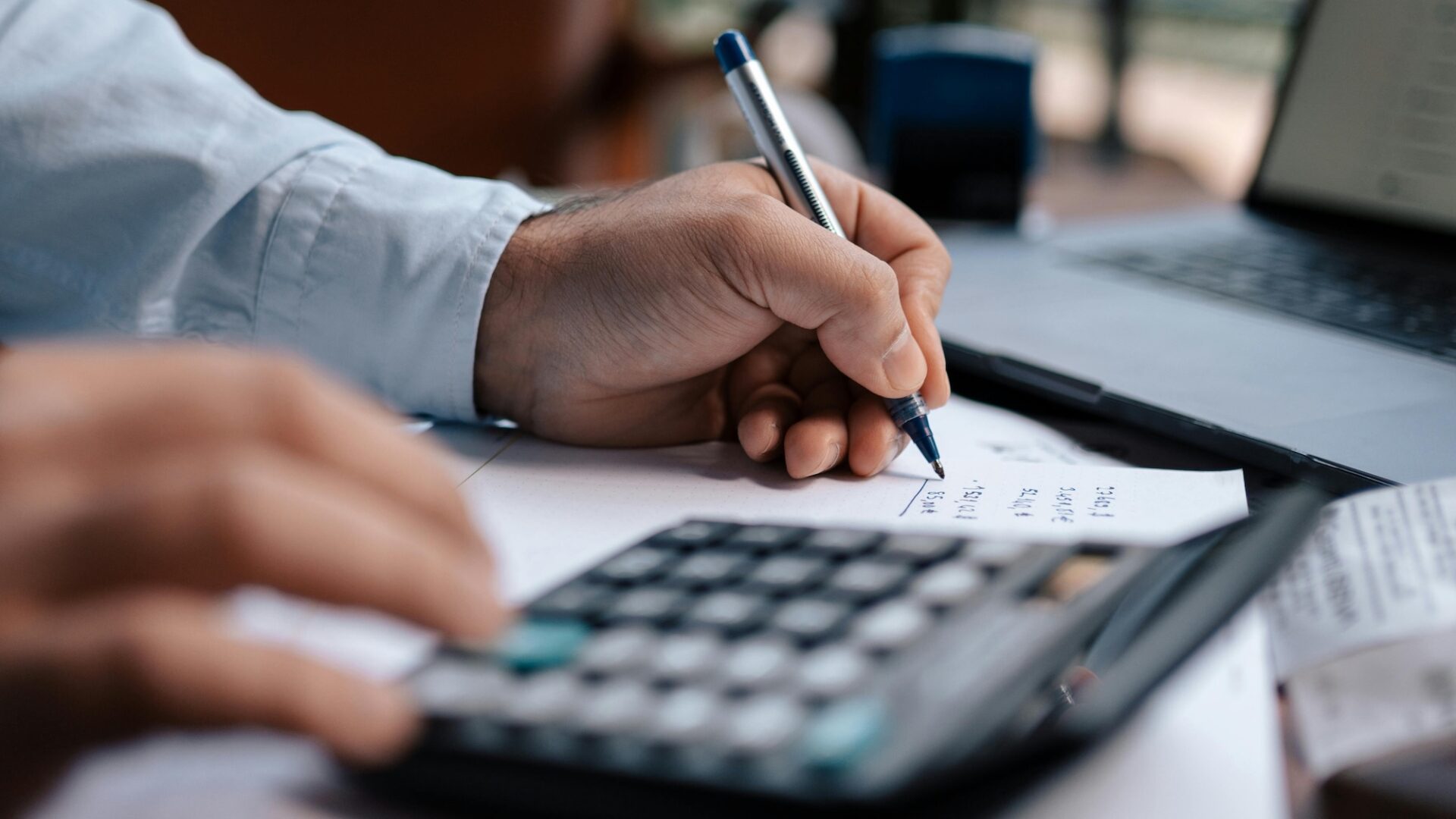 A real estate agent uses a calculator, laptop and pen and paper to figure out their email marketing costs