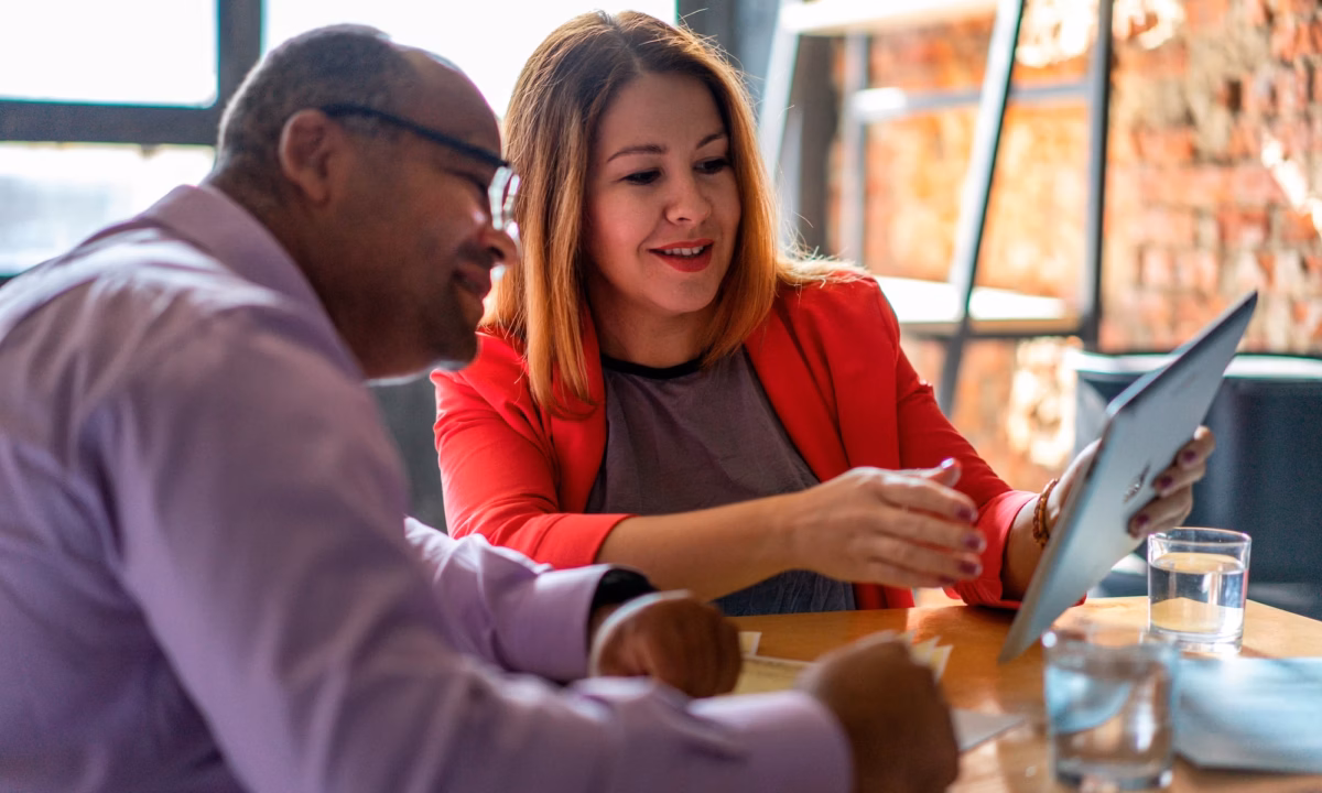 An agent sits with a client at a desk, showing him a real estate listing on her white-labeled mobile app